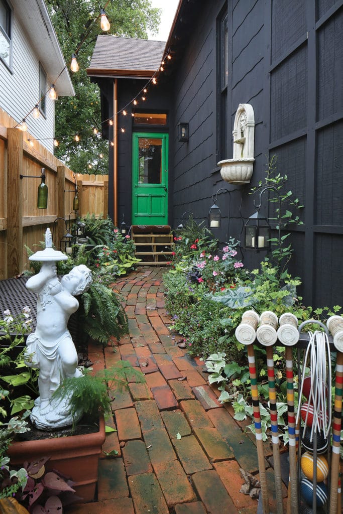 Courtyard of Cottage or bungalow style home painted black. This photo features a bright green door and lots of flowers in bloom.