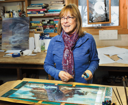 A woman stands at a work table with materials used in stained-glass making.