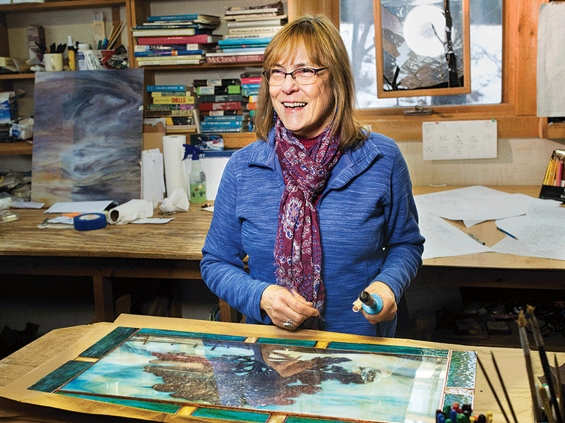 A woman stands at a work table with materials used in stained-glass making.