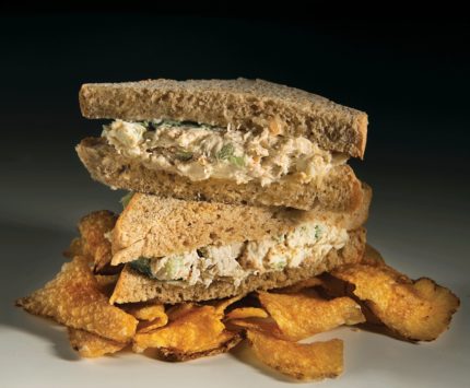 A chicken salad sandwich on wheat bread sits atop potato chips.