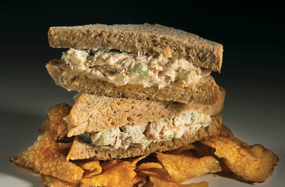 A chicken salad sandwich on wheat bread sits atop potato chips.