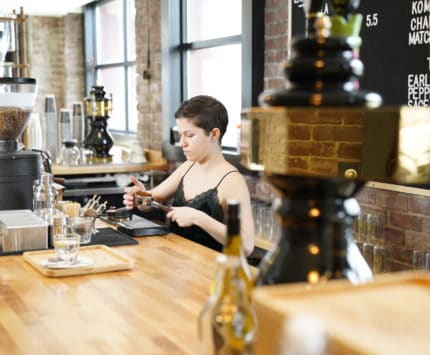 A woman prepares coffee behind the high wooden counter at a barbershop.