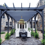 A dark stained pergola with a large dining table sits outside.