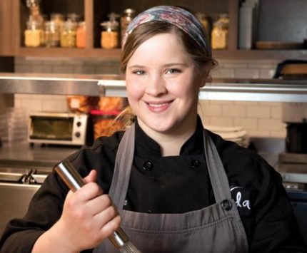 A female chef stands smiling in a kitchen while whisking something in a metal bowl.