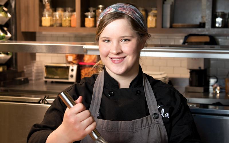 A female chef stands smiling in a kitchen while whisking something in a metal bowl.