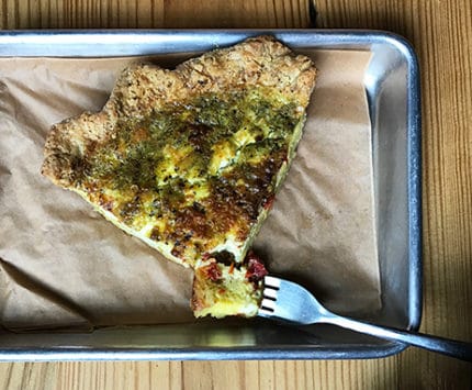 A silver tray holds a giant slice of green pesto queiche.