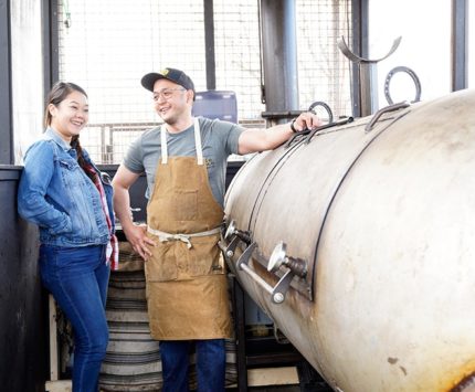 A woman and man stand next to a barbecue smoker.