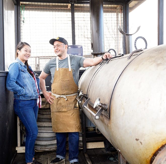 A woman and man stand next to a barbecue smoker.