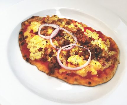 A large, golden brown slab of flatbread holds yellow puffs of eggs, chorizo sausage, and sliced red onion.