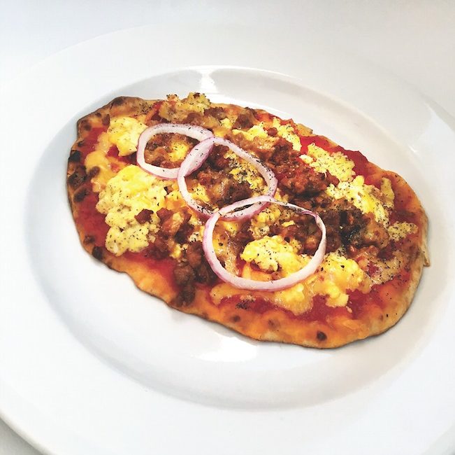 A large, golden brown slab of flatbread holds yellow puffs of eggs, chorizo sausage, and sliced red onion.