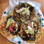 A paper plate with three tacos topped with salsa, cabbage, sour cream, and avocados