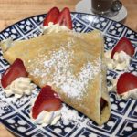 A plate of a sweet crepe filled with Nutella and surrounded by whipped cream and strawberries