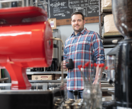 A tall man holding a coffee mug stands behind a large, bright red, industrial coffee grinder.