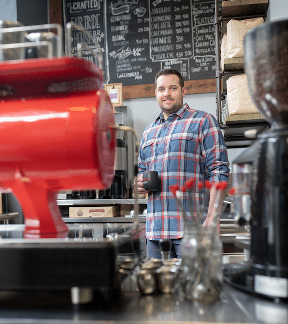 A tall man holding a coffee mug stands behind a large, bright red, industrial coffee grinder.