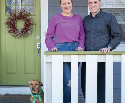 A woman and a man stand on the front porch of a house.