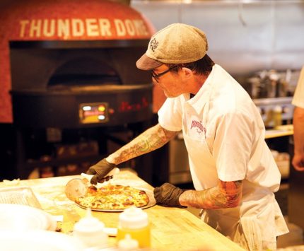 A tattooed man slices into a pie that's just been plucked from a pizza oven.