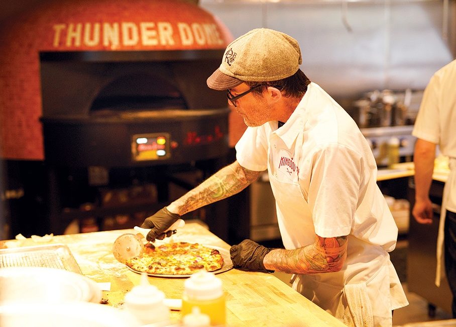 A tattooed man slices into a pie that's just been plucked from a pizza oven.