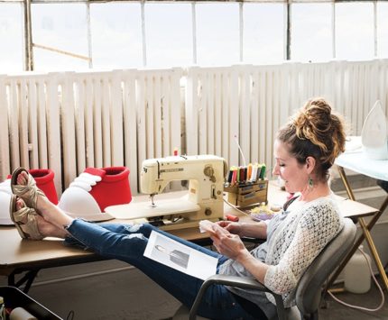 A woman sits with her feet up at a sewing machine desk while stitching together a doll.