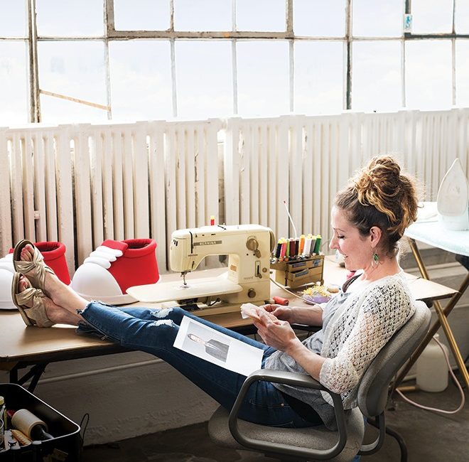 A woman sits with her feet up at a sewing machine desk while stitching together a doll.