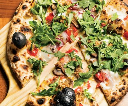 A perfectly charred pizza sits atop a pizza paddle and is dressed with arugula.