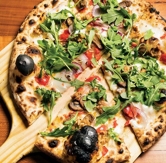 A perfectly charred pizza sits atop a pizza paddle and is dressed with arugula.