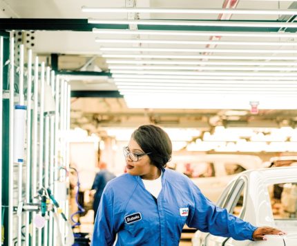 A woman dons a blue work jumpsuit and safety glasses while working in a car factory.