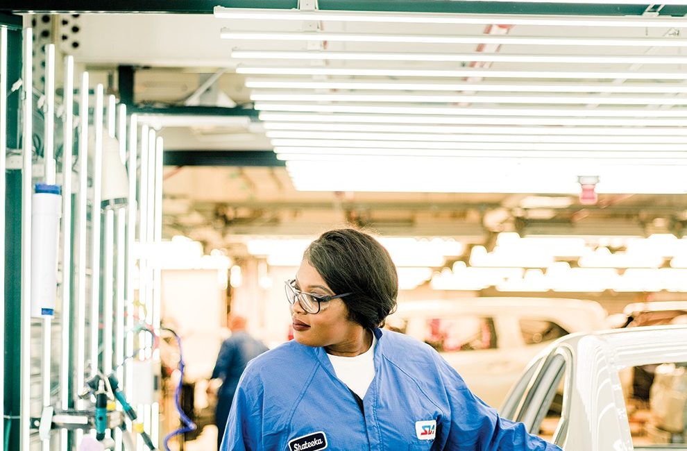 A woman dons a blue work jumpsuit and safety glasses while working in a car factory.