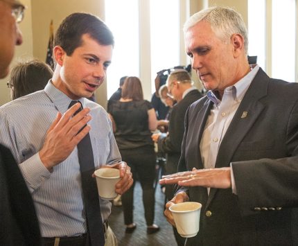 Pete Buttigieg and Mike Pence have a conversation while gesturing with their hands and holding coffee mugs.