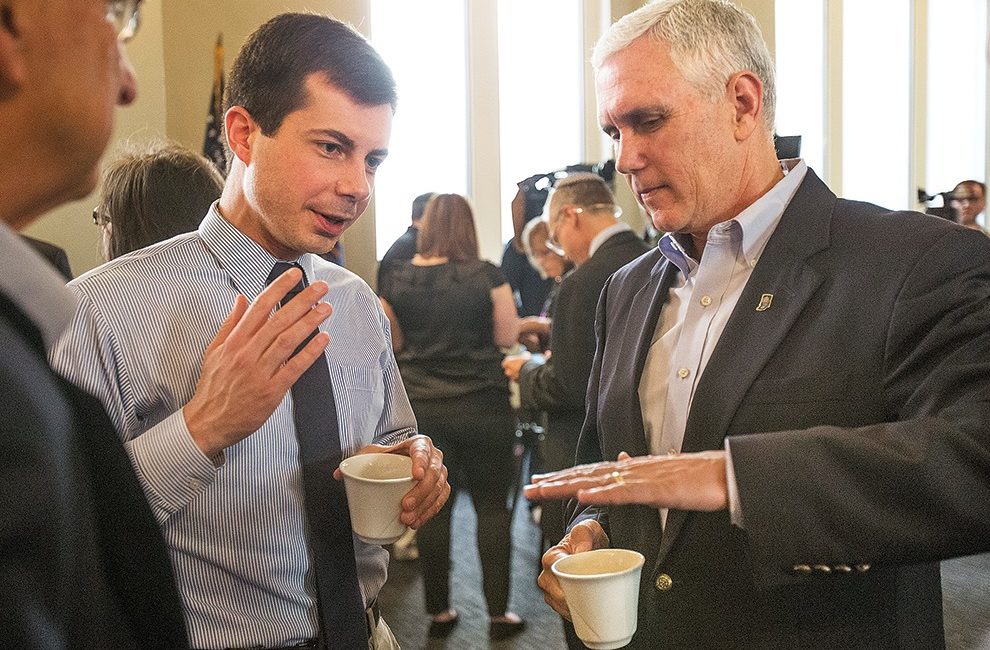 Pete Buttigieg and Mike Pence have a conversation while gesturing with their hands and holding coffee mugs.