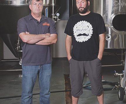 Two men stand before brewing vats.