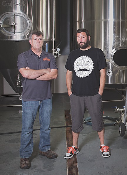 Two men stand before brewing vats.