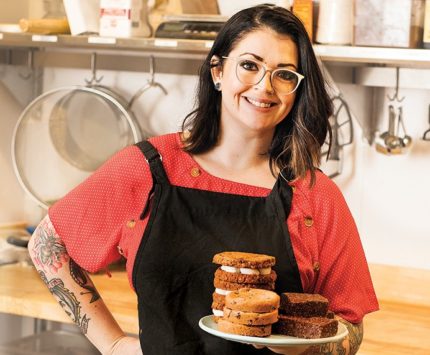 A woman dons a black apron while holding a plate of pastries.