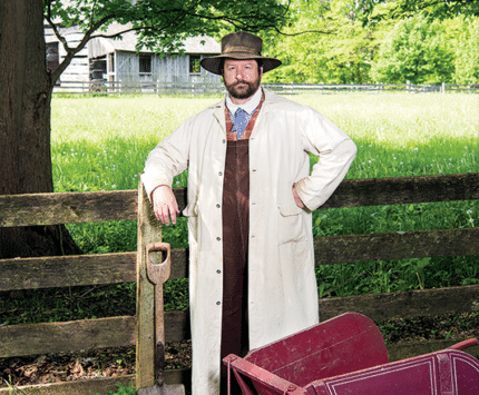 A historical interpreter stands along a fence post, dressed in 19th century garb.