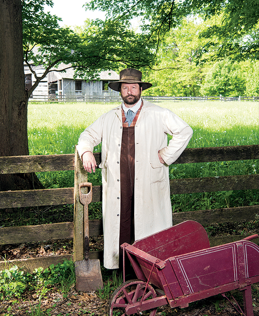 A historical interpreter stands along a fence post, dressed in 19th century garb.