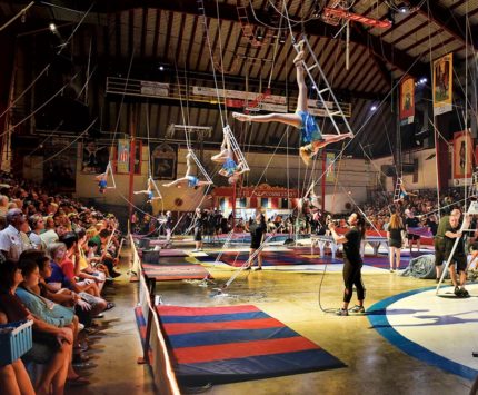 Colorful circus demonstration with trapeze artists dangling from ropes high above the ground.