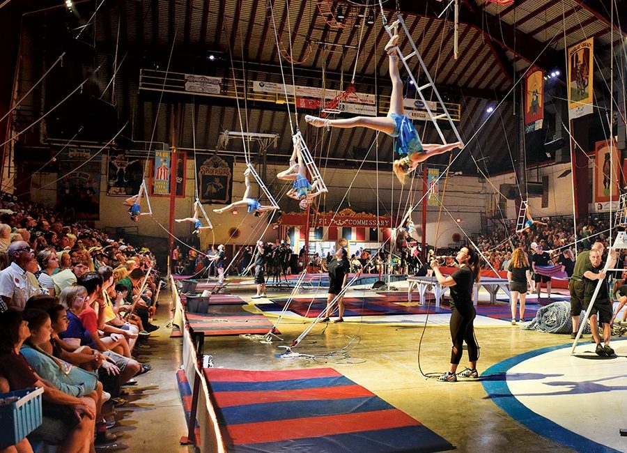 Colorful circus demonstration with trapeze artists dangling from ropes high above the ground.