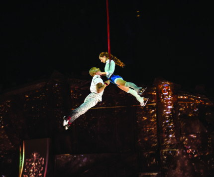 Two trapeze artists dangle midair at a stage performance.