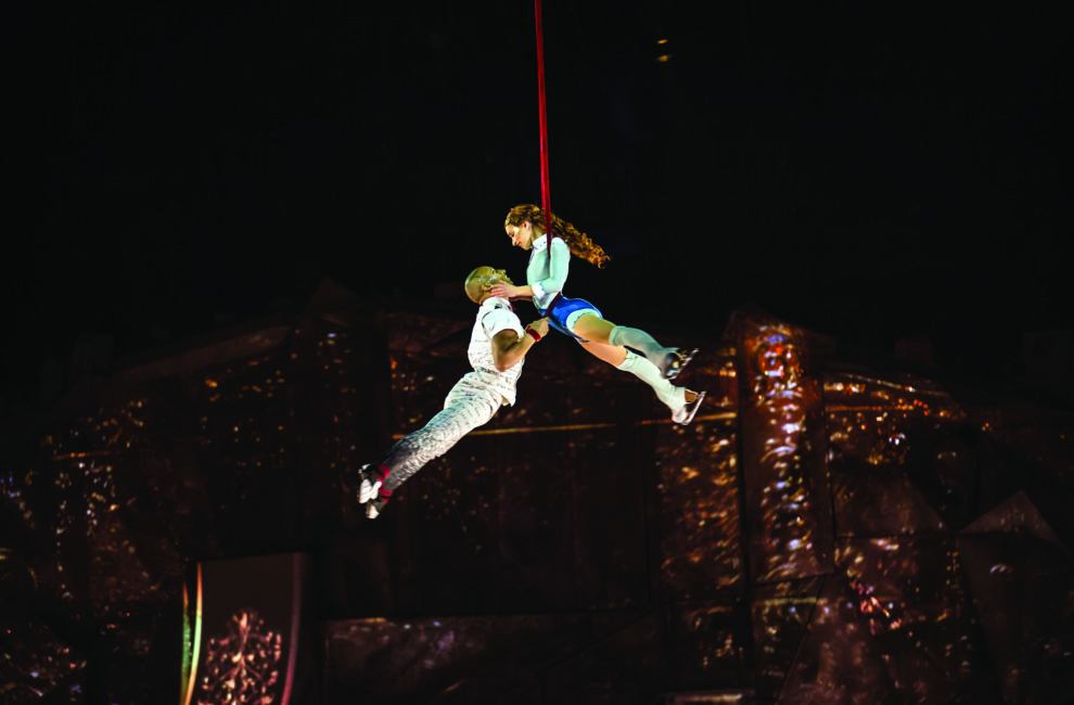 Two trapeze artists dangle midair at a stage performance.