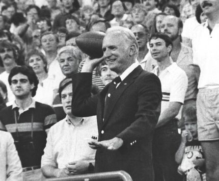 Historic black and white photo of a man throwing a football from the stands to begin an NFL game.