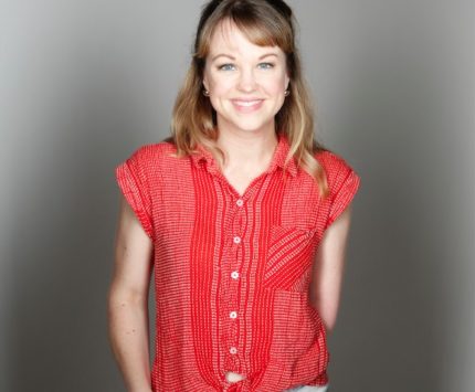 A woman stands against a gray studio background wearing a red shirt tied at the waist and light wash jeans.