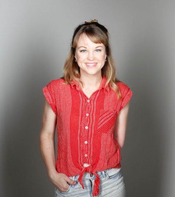 A woman stands against a gray studio background wearing a red shirt tied at the waist and light wash jeans.