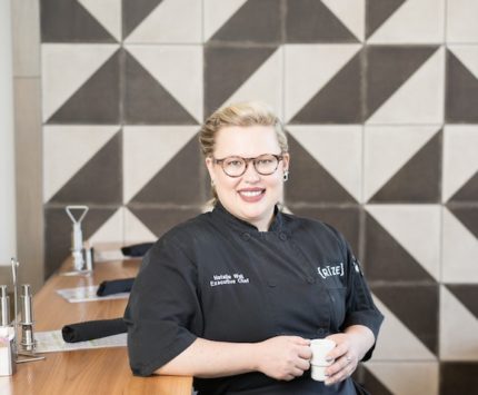 A woman in a chef’s apron sips coffee against a black and white geometric wall.