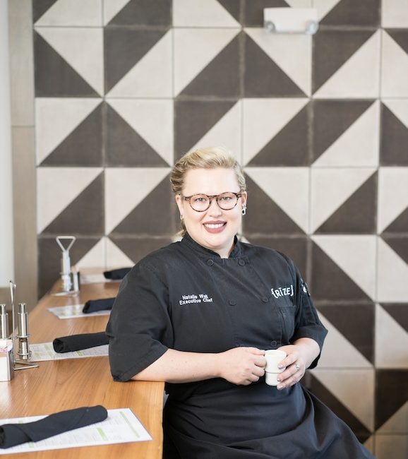 A woman in a chef’s apron sips coffee against a black and white geometric wall.