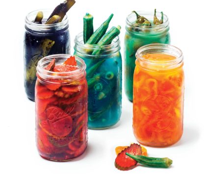 Five brightly-colored mason jars containing different types of pickles in Kool-Aid