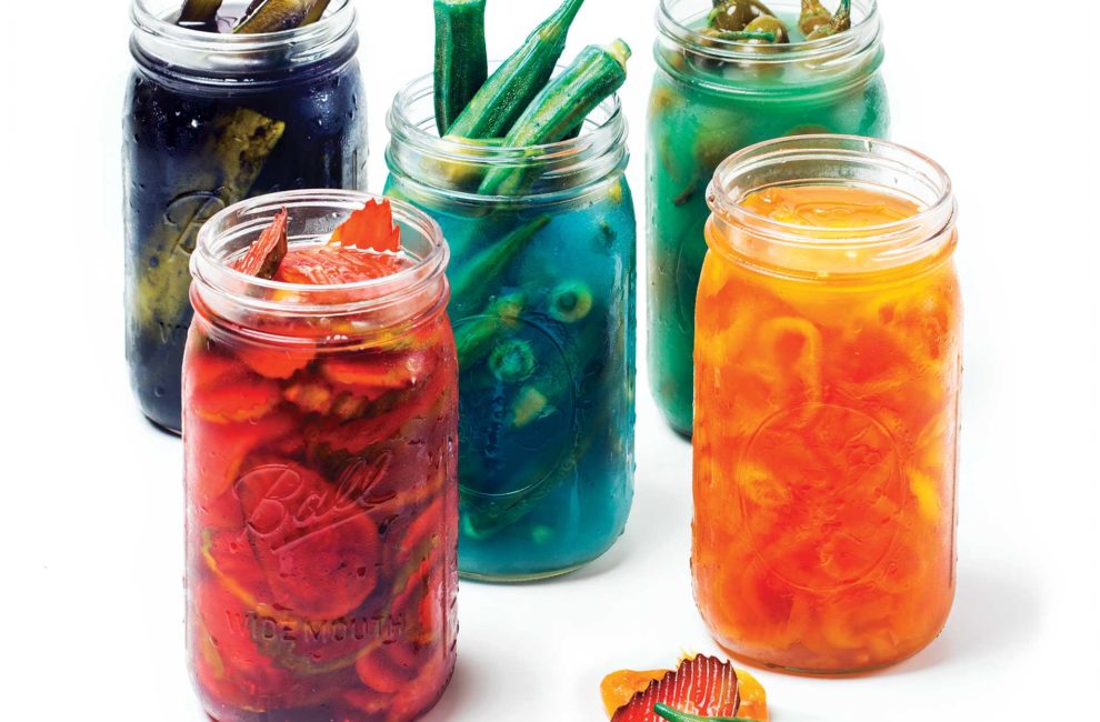 Five brightly-colored mason jars containing different types of pickles in Kool-Aid