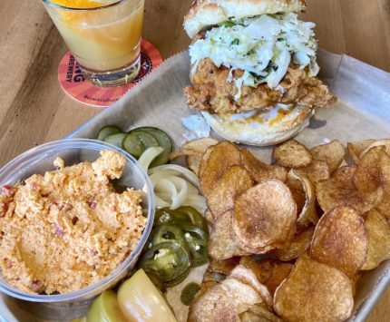 A tray of food featuring a chicken sandwich, potato chips, and pimento cheese, with a cocktail.