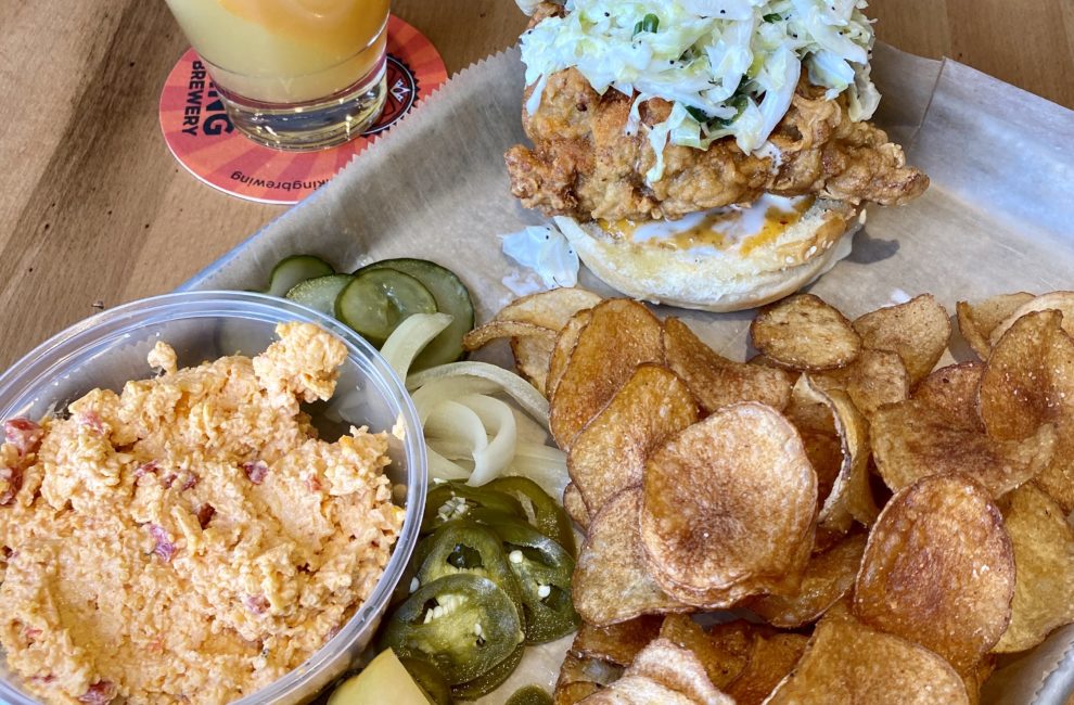 A tray of food featuring a chicken sandwich, potato chips, and pimento cheese, with a cocktail.