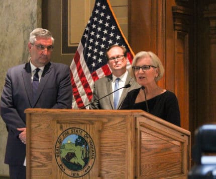 Dr. Kristina Box stands at a podium while Indiana Governor Eric Holcomb waits by her side.