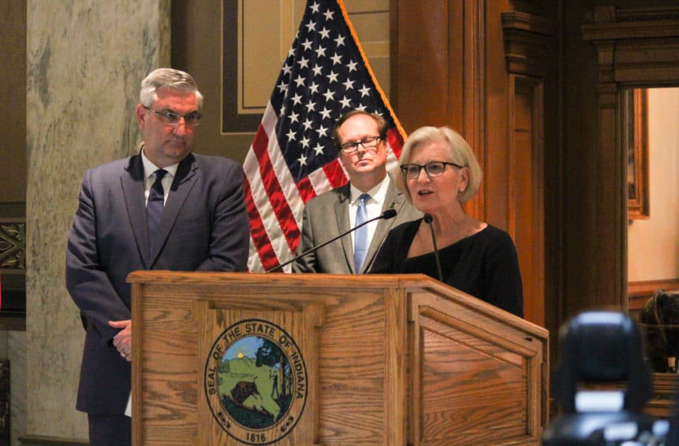 Dr. Kristina Box stands at a podium while Indiana Governor Eric Holcomb waits by her side.