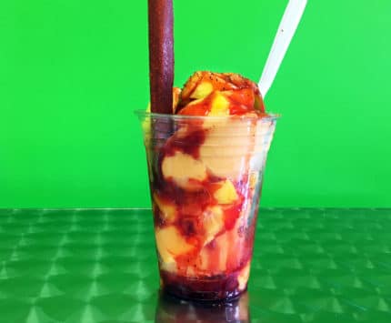 Against a green background, a plastic parfait cup is filled with scoops of mango ice cream with reddish brown chili syrup, a white spoon, and a tamarind candy stick